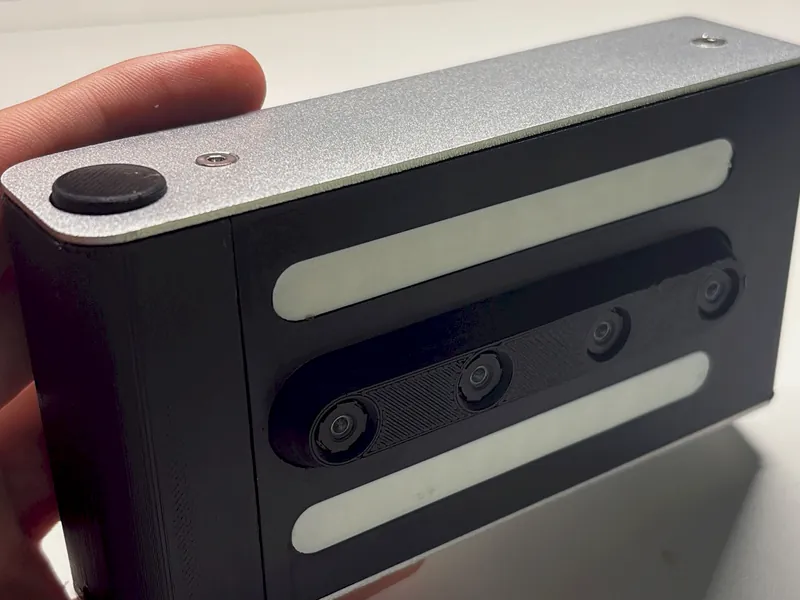 The black merged. prototype held at a desk: aluminium top plate, shutter button, two white flash bars around the four-lens strip.