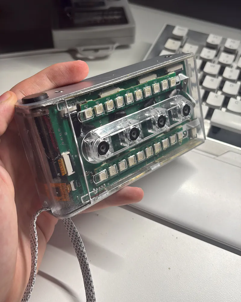 An early merged. prototype in a clear housing, held in hand at a desk: green circuit boards, LED rows and four lenses visible through the case.