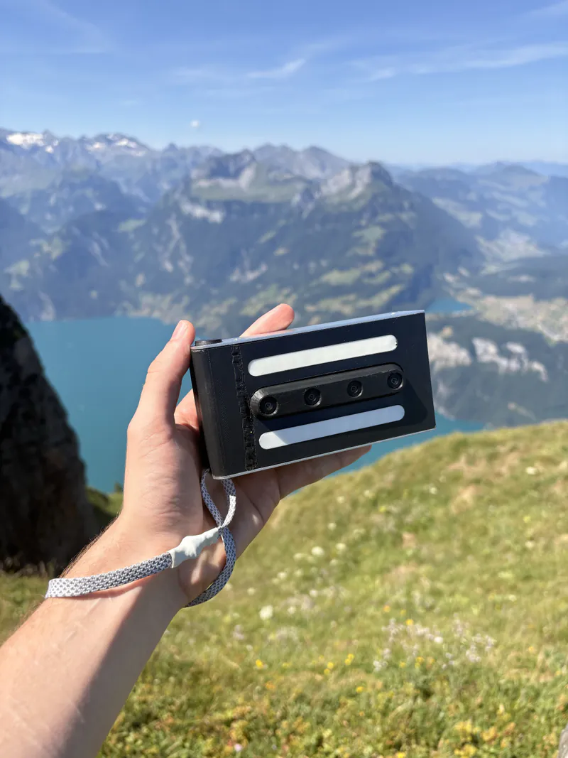 A hand holds the black merged. prototype, its four lenses in a horizontal row, against an alpine lake and mountain range on a clear day.